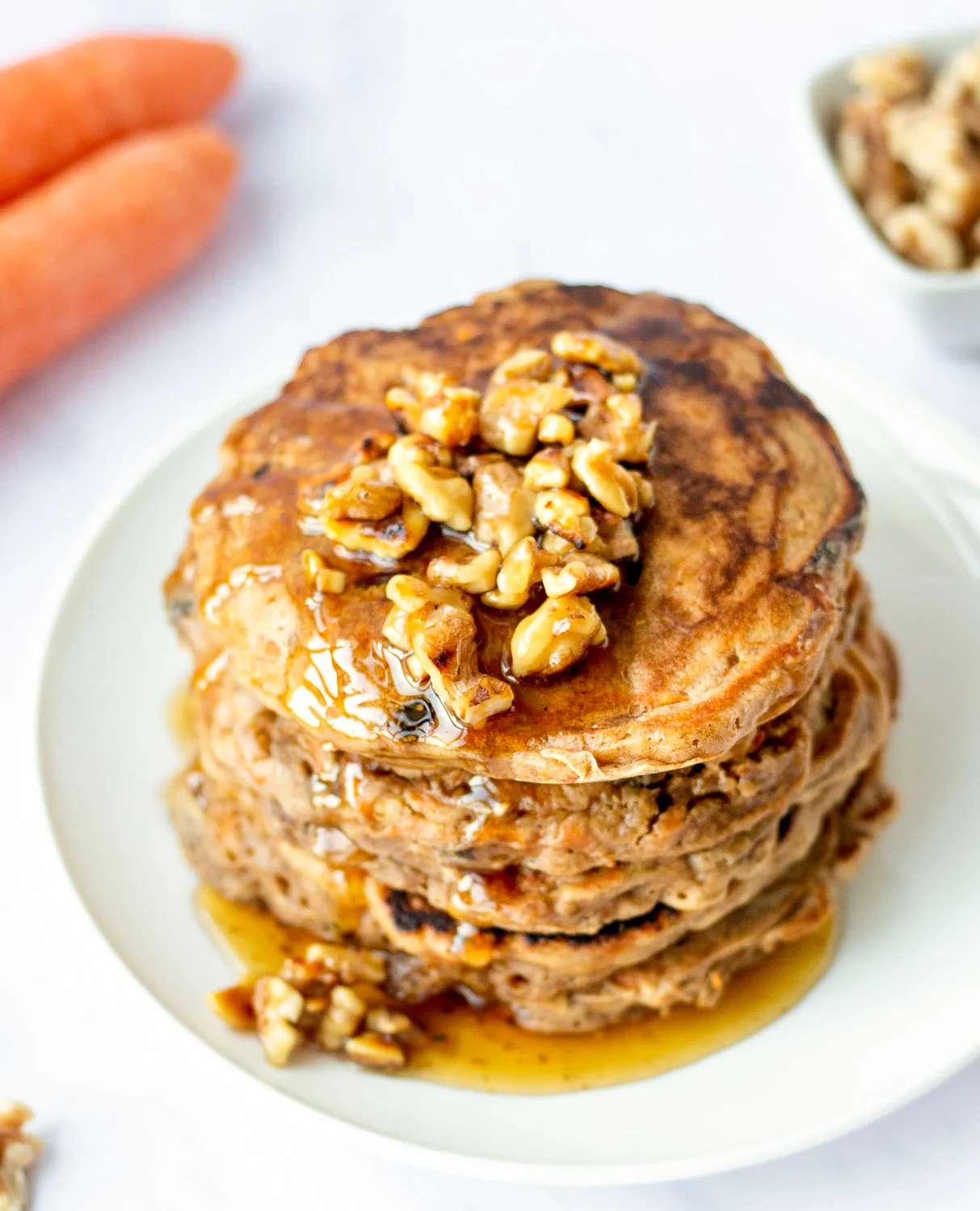 stack of carrot cake pancakes topped with walnut syrup and a carrot in the background