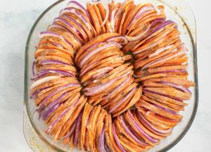 Thinly sliced sweet potatoes stacked vertically in a roasting pan with red onion slices in between.