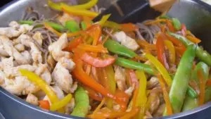 stir frying soba noodles, chicken, and veggies in peanut sauce in a pan