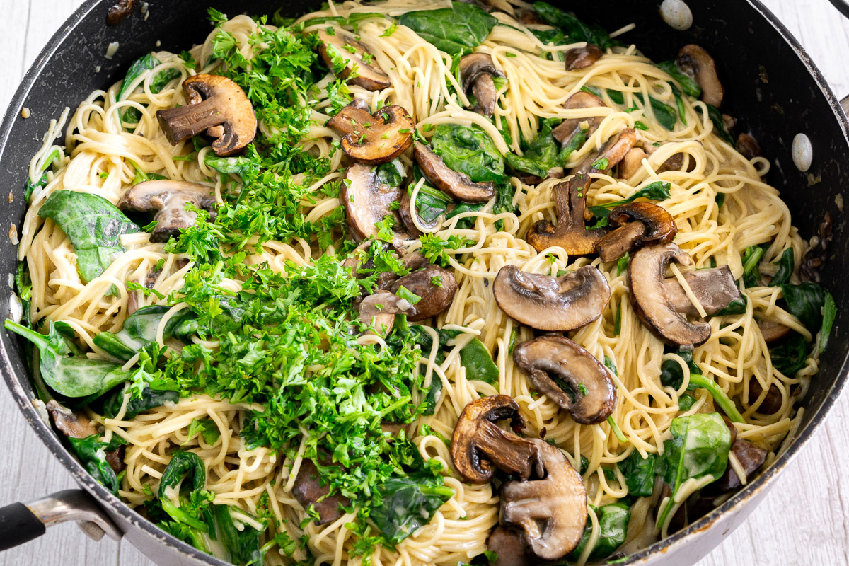 close up of angel hair pasta, mushrooms, spinach with creamy sauce and herbs in a pan