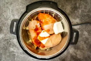 photo of Instant Pot with ingredients for Buffalo chicken inside: chicken, Buffalo sauce, cream cheese, and onions