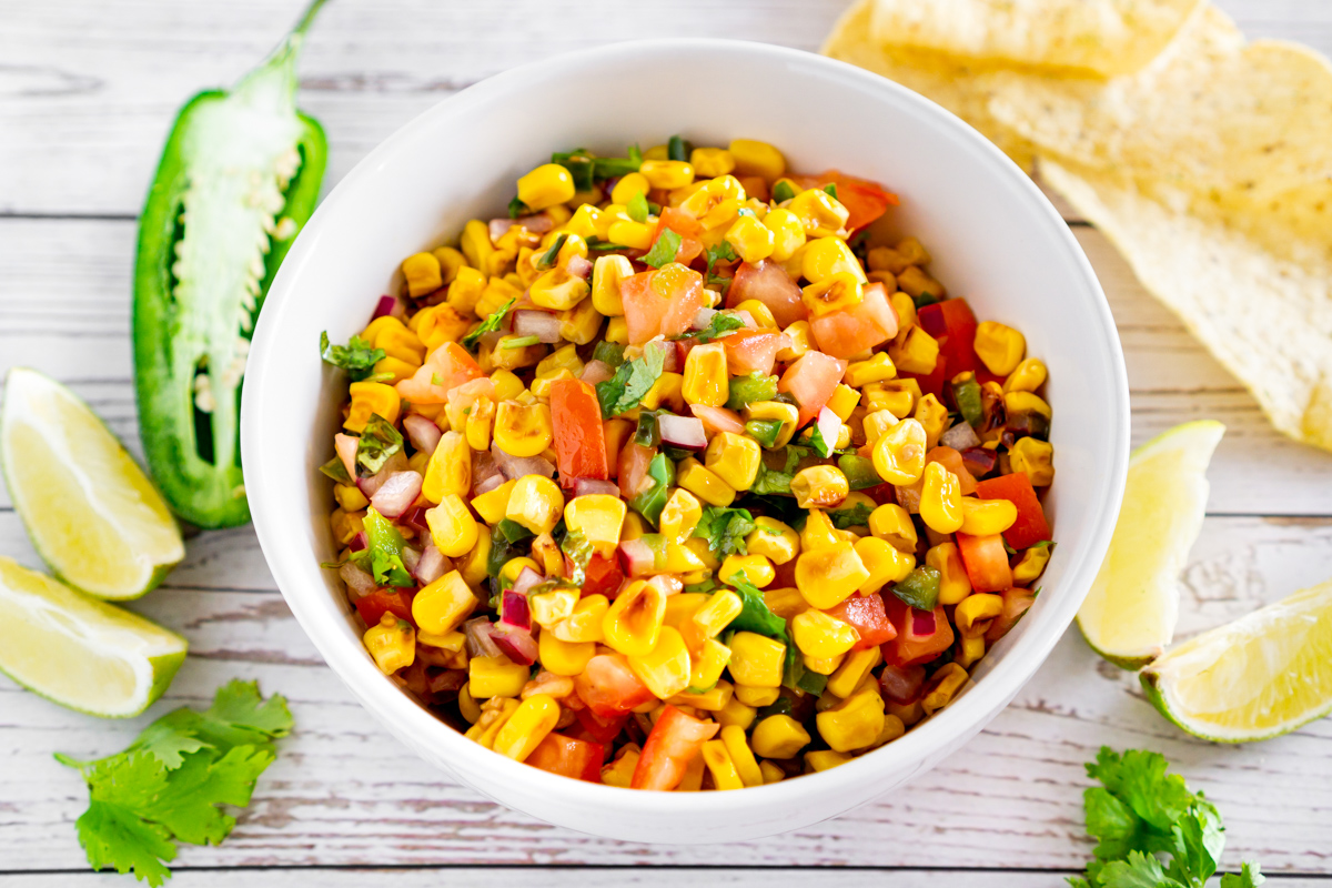 charred corn salad in a bowl surrounded by jalapeno, lime, chips