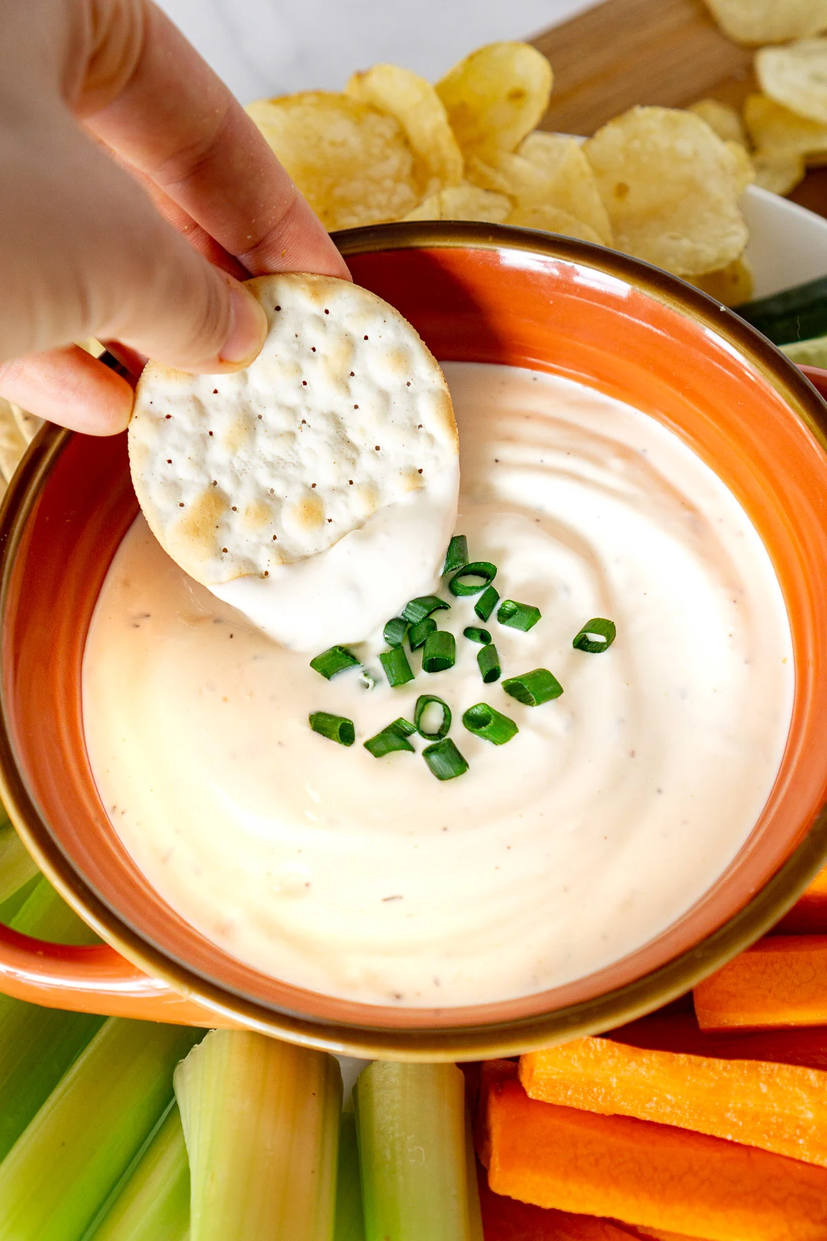 hand dipping cracker into a creamy dip with veggies on the side