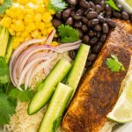 Bowl of blackened salmon on rice with avocado, black beans, corn, cucumber, red onion.