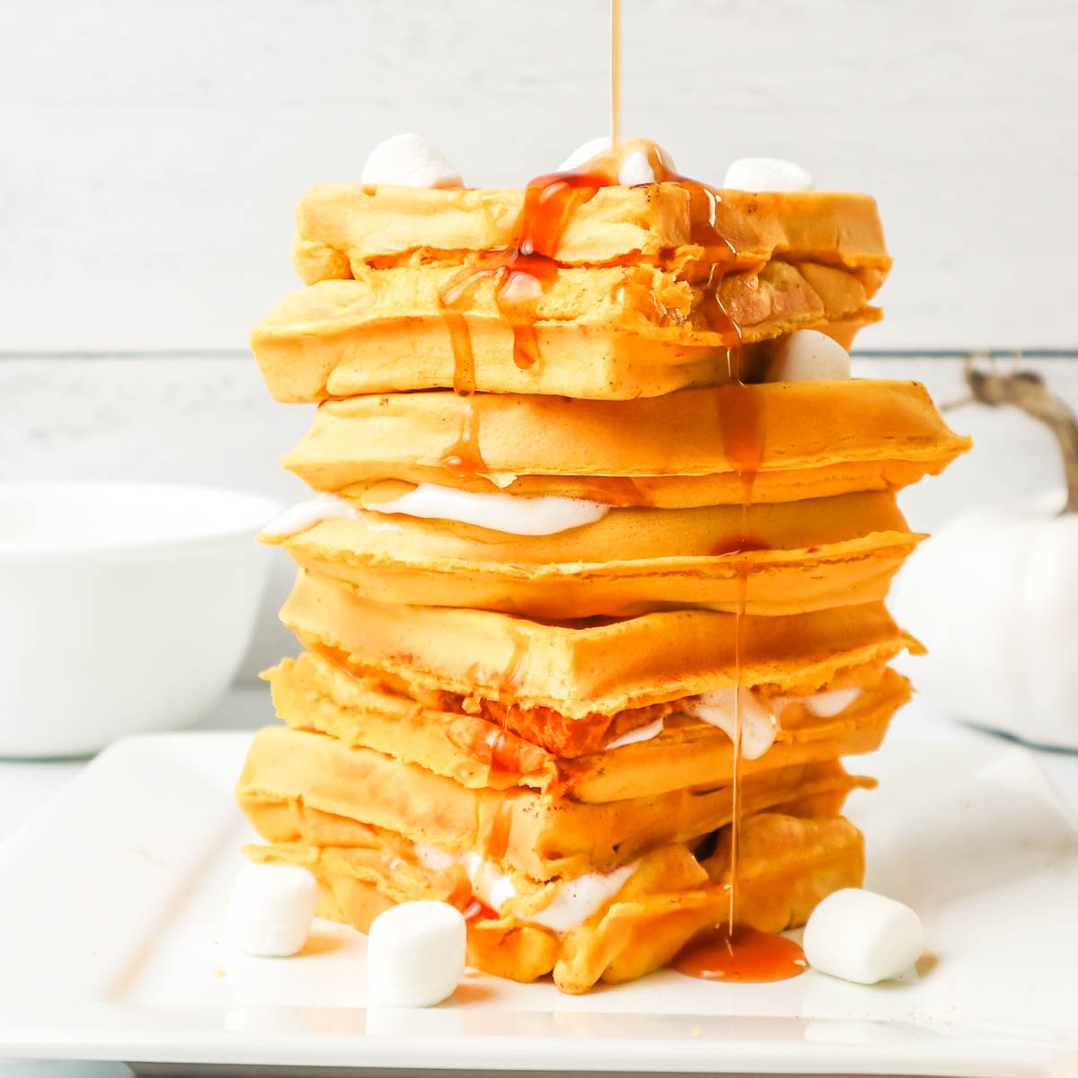 Stack of pumpkin waffles with marshmallows with syrup being poured on top.