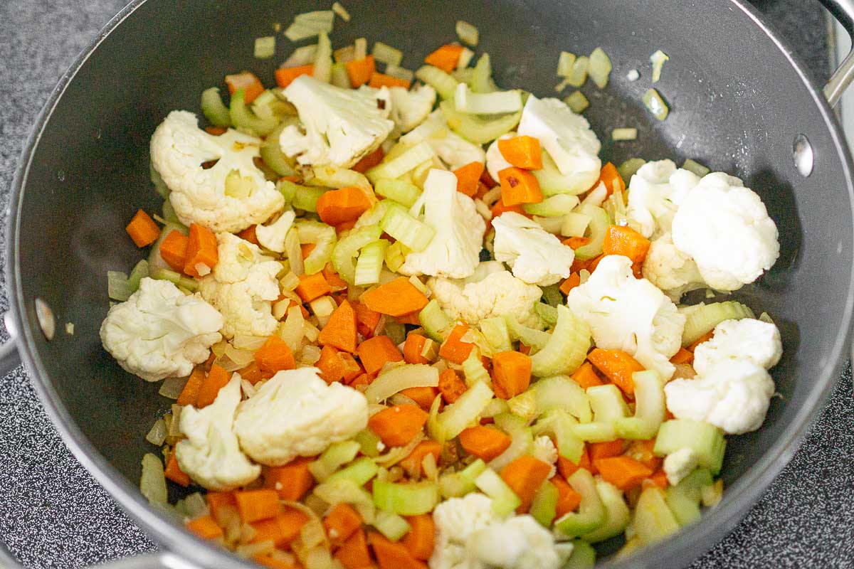 Adding cauliflower and celery to veggies in soup pot.