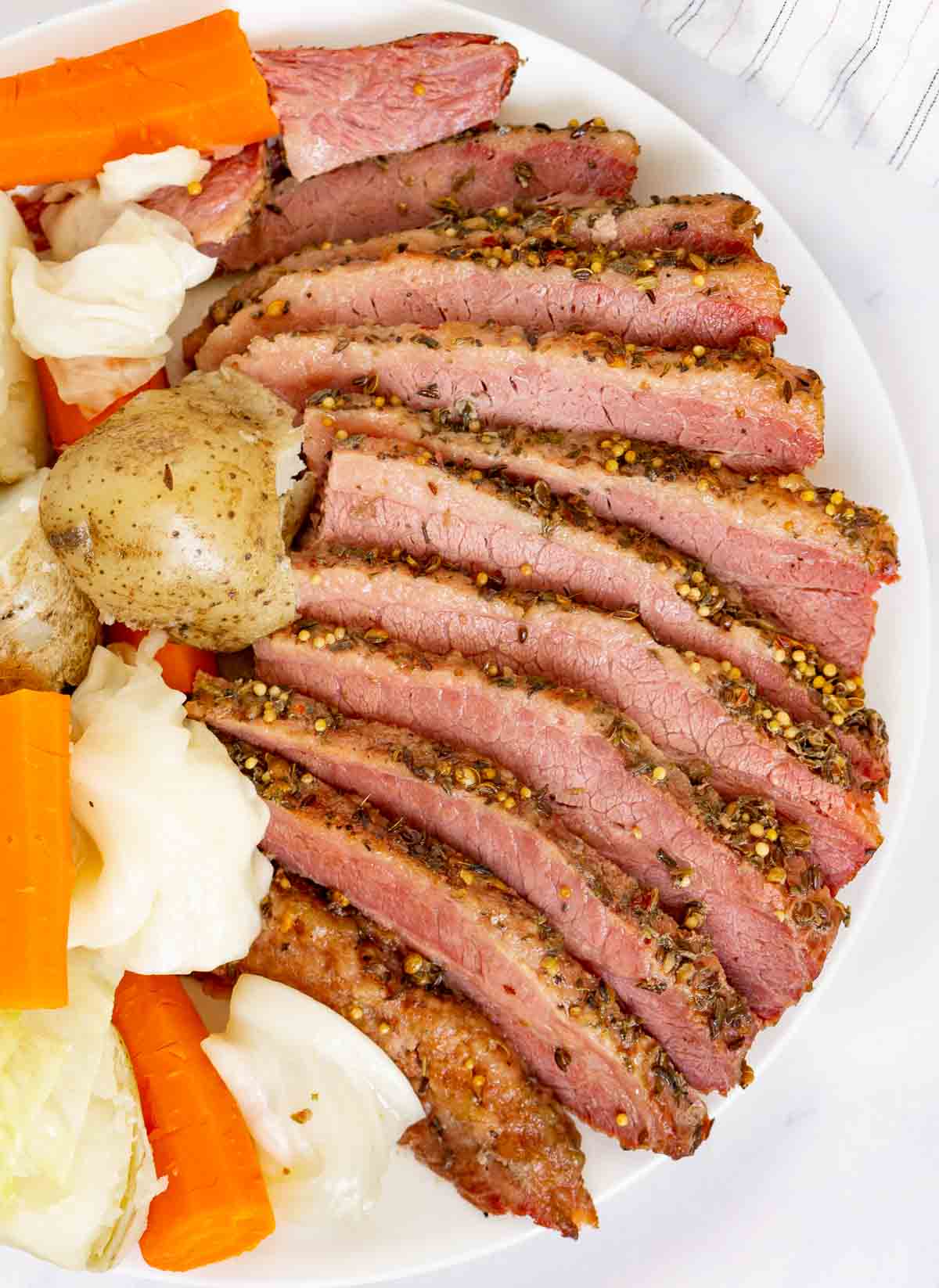 Sliced corned beef on a plate next to potatoes, cabbage, and carrots.