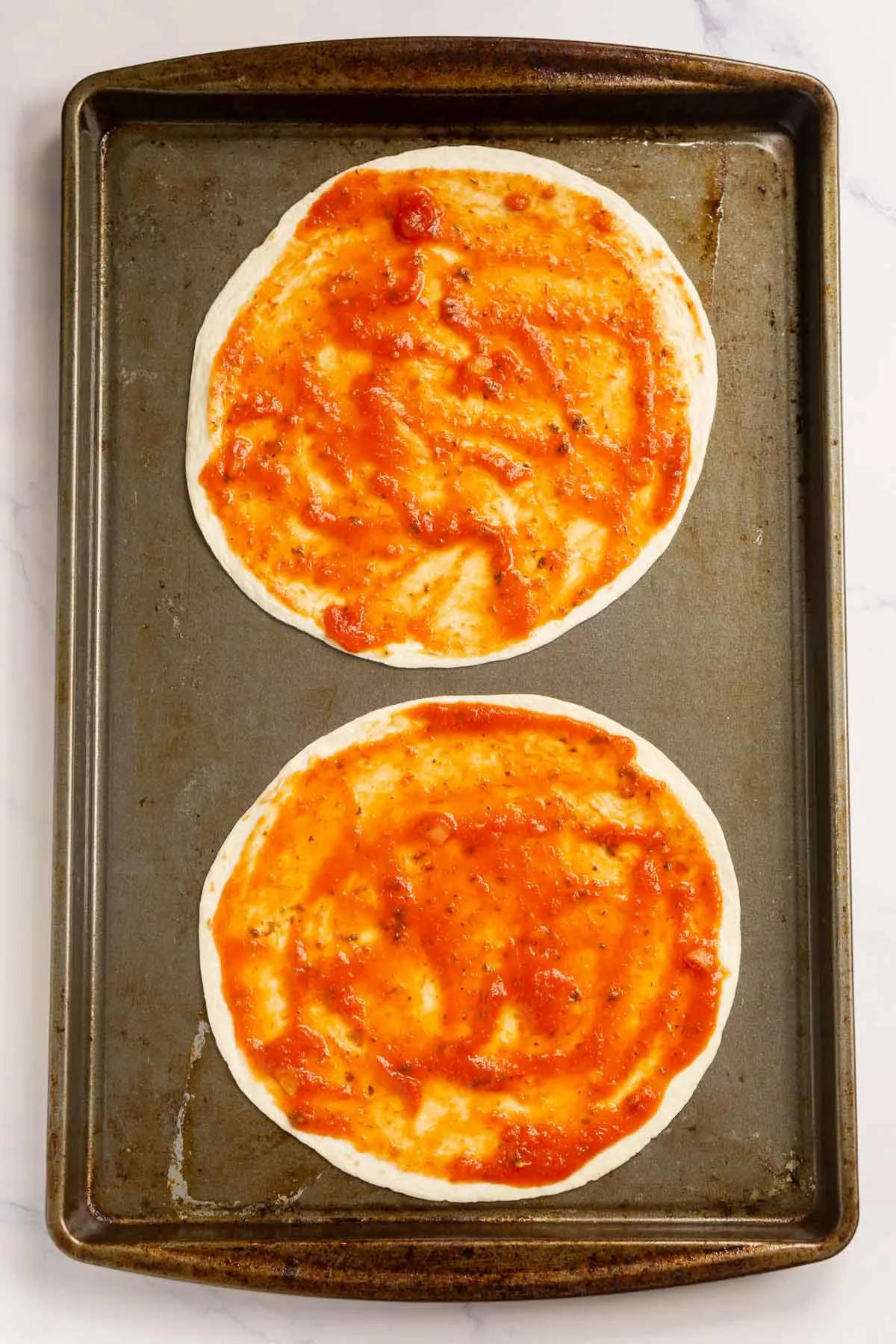 Two tortillas on a baking sheet with pizza sauce spread on them.