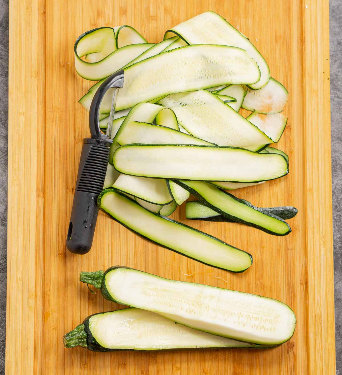 Sliced zucchini ribbons on a cutting board next to a vegetable peeler